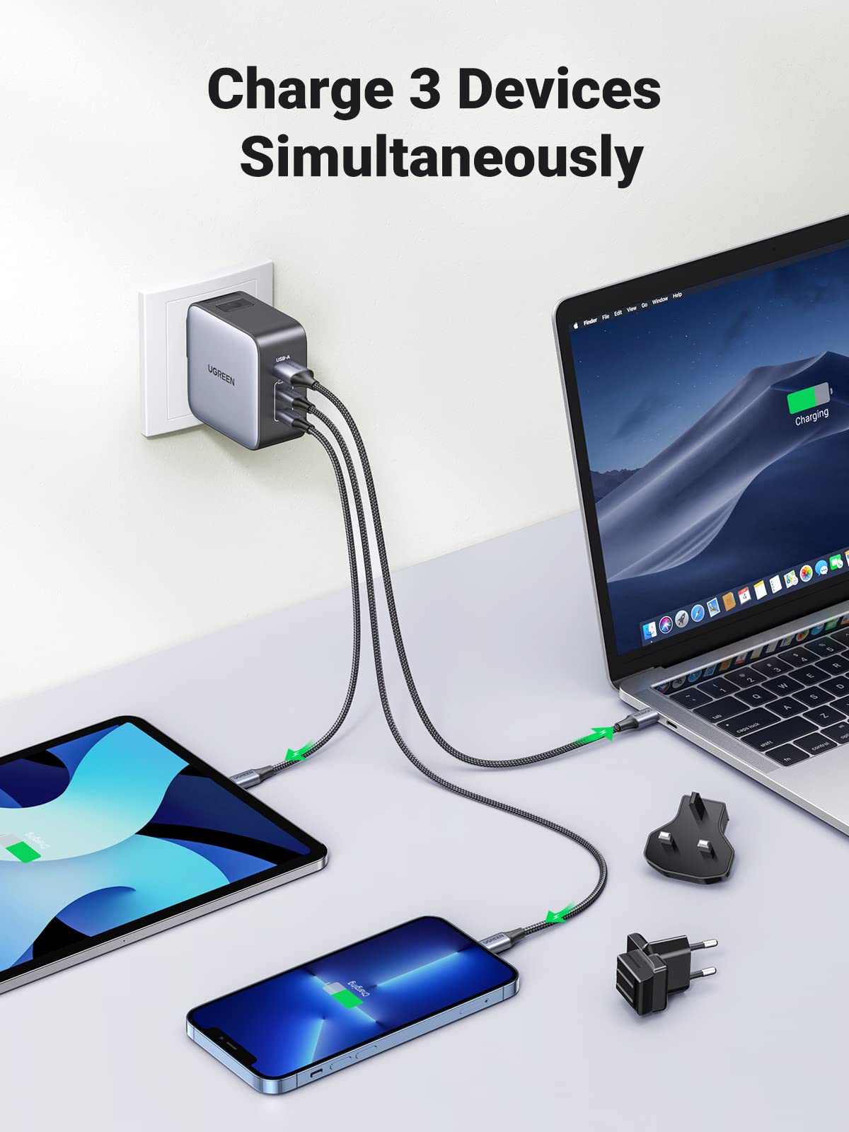 UGREEN 65W iphone charger's ability to charge three devices simultaneously. It the charger plugged into a wall socket, with cables connected to a laptop, a tablet, and a smartphone, all indicating they are charging. Two additional plug adapters are also visible on the table.